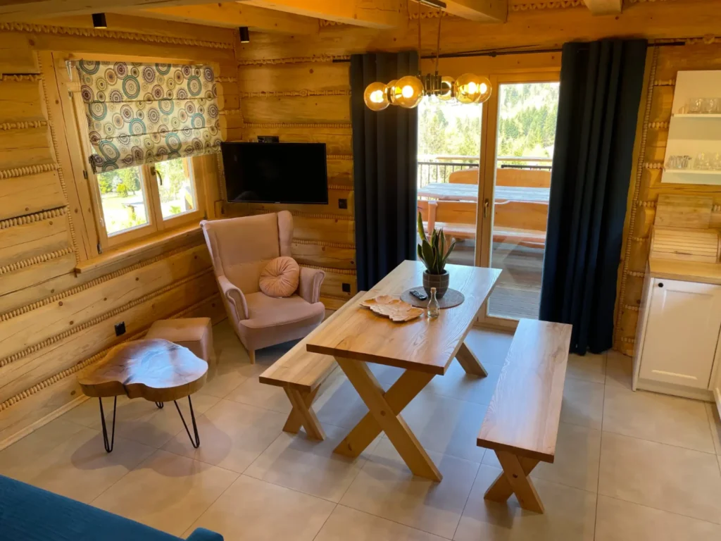 jadalnia, duzy stół drewniany z dwoma ławkami na środku, okno z widokiem na balkon, fotel z podnóżkiem, telewizor