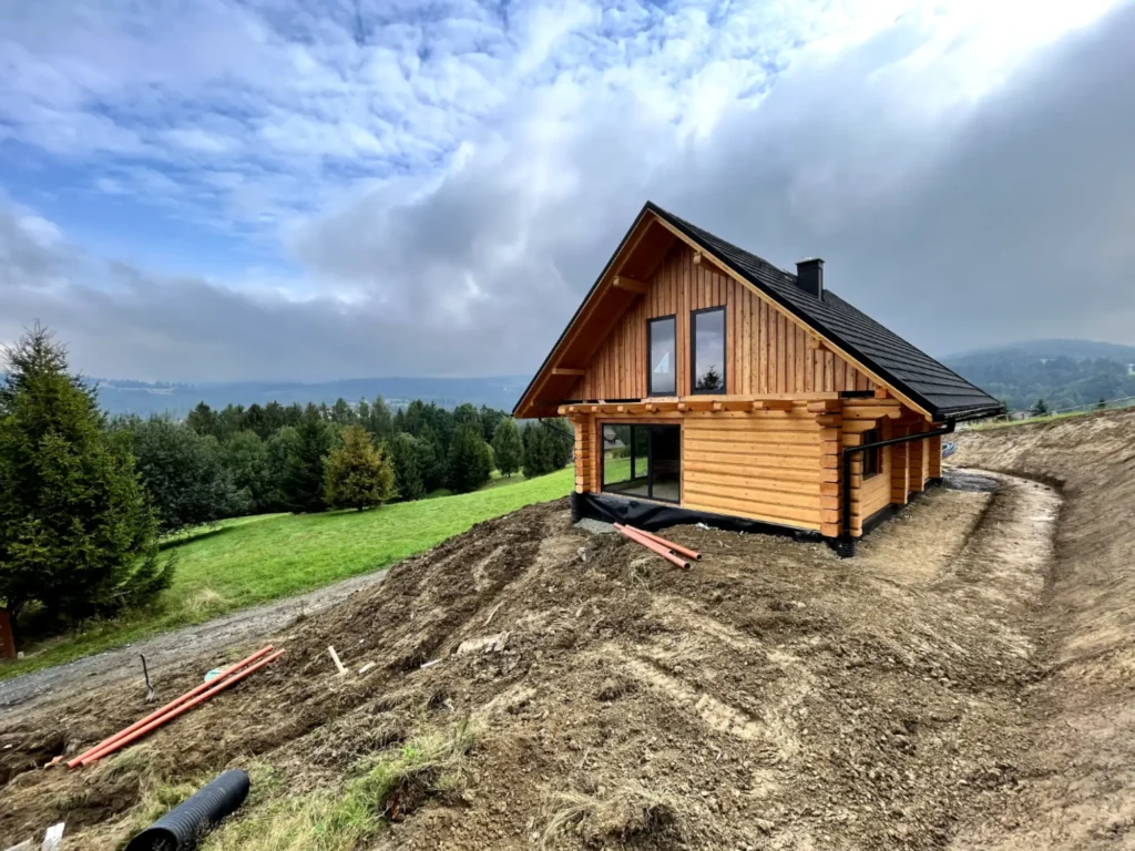 dom z bali na wzgórzu, kuchnia z dużym oknem, dach dwuspadowy, widok na okolicę, łąki i lasy, pochmurna pogoda